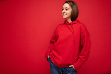 Photo shot of charming positive smiling brunette female teenager wearing stylish red hoodie standing isolated on red background wall looking to the side