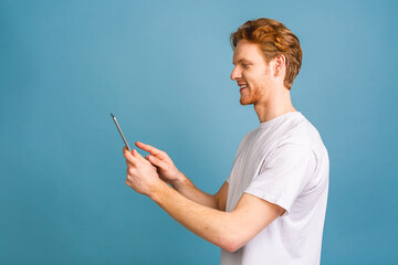 Happy young bearded red-haired man in casual using tablet and standing isolated over blue background.
