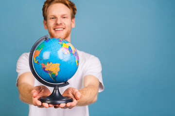 Portrait of young bearded businessman wants to explore unknown places, looks at worldwide globe, tries to choose destination, isolated over blue background. Young male holds earth indoor