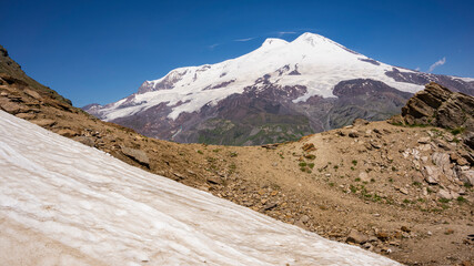 Volcano Elbrus is dormant volcano located in the south-east Caucasus mountain range in Kabardino-Balkaria, Russia. View from Cheget mount slope. There is snow in the foreground. Altitude is 3050 m.