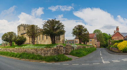 Ancient All Saints Church flanked by dry stone walls under blue sky. Goodmanham, UK.