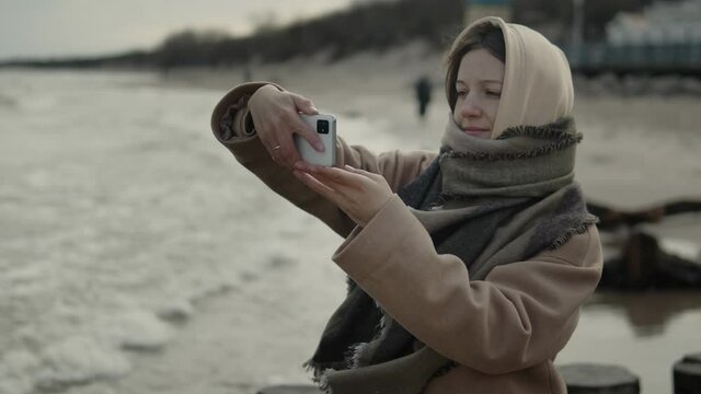 Young woman wearing outwear takes photos with her smartphone on the beach by the sea. Girl in warm coat with hood on her head walking on empty beach