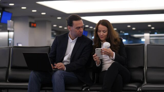 Woman ask man to look at smartphone, he turn away from laptop and watch to phone. Two people wait for boarding at modern domestic terminal. Formal smart casual clothes style