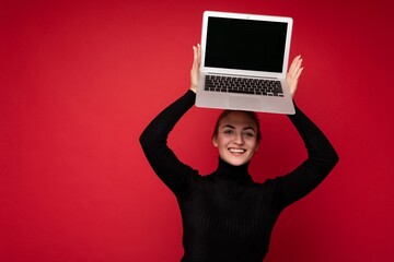 Photo of Beautiful smiling happy young brunet lady holding computer laptop with empty monitor screen wearing black longsleeve looking at camera isolated over red wall background