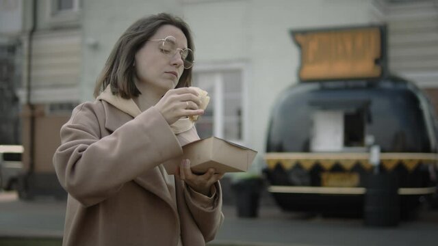 Close up of a millenial woman in round glasses and oversized beige coat eating street food from cardboard container outdoors. Foodtruck at the background. Brunette woman having a lunch in the streets