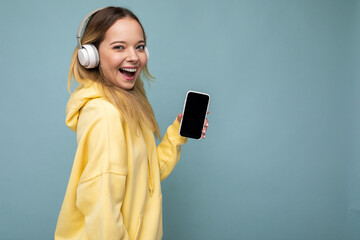 Side-profile photo of beautiful happy smiling young woman wearing stylish casual outfit isolated on background wall holding and showing mobile phone with empty display for mockup wearing white