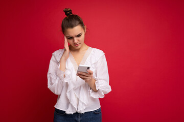 Attractive young upset brunet woman wearing white blouse standing isolated over red background reading news on the internet via phone looking at mobile screen