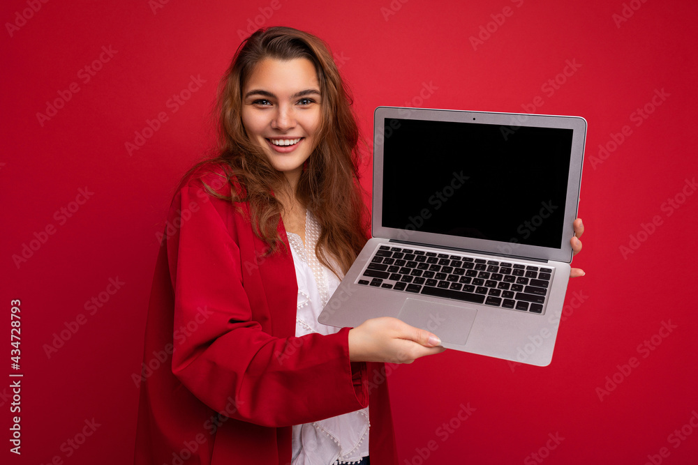 Wall mural Close-up portrait of Beautiful smiling happy young brunette woman holding computer laptop with empty monitor screen wearing red cardigan and white blouse looking at camera isolated over red background