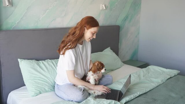 A pregnant woman working at home on a laptop sits on the bed in the bedroom and holds her pet spaniel in her arms