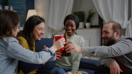 Afro american woman talking with her friends holding beer bottle while sitting on sofa late at night in living room. Group of mixed race people hanging out enjoying time spending together