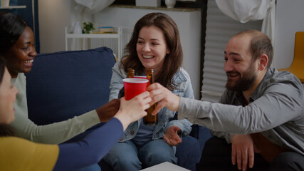 Group of multi-ethnic happy friends having fun during entertainment wekeend party drinking beer, eating snack while relaxing on sofa. Mixed race people hanging out enjoying time spending together