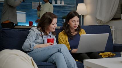 Group of multiethnic people discussing while watching entertainment video on laptop resting on couch in living room. In background afro american woman and man drinking beer enjoying free time together