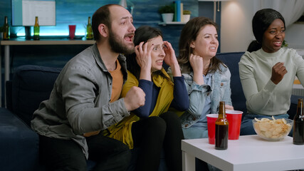 Group of mixed people watching sport on television at home cheering football team. Multi-ethnic friends sitting on couch celebrating soccer goal having fun together late at night