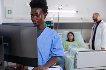 Black medical asisstant typing sickness treatment on computer while physician doctors monitoring sick woman analyzing diagnosis. Patient lying in hospital ward bed during consultation recovery