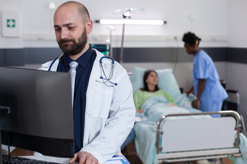 Front view of practitioner doctor typing medical expertise on computer while in background black asisstant discussing healthcare treatment. Hospitalized patient having respiratory disorder