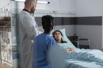 Sick woman patient discussing with medical doctors while resting in bed during illness recovering in hospital ward. Man practitioner doctor writing sickness recovery on clipboard