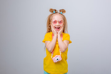 little girl in sunglasses traveler holds a camera on a white background. Funny cute little girl in a yellow T-shirt and sunglasses smiling on a white background, emotional