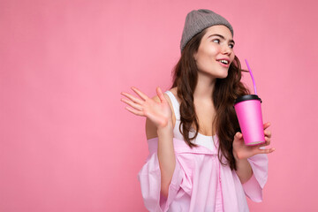 Charming young happy brunette woman wearing stylish clothes isolated over colourful background wall holding paper cup for mockup drinking milkshake looking to the side