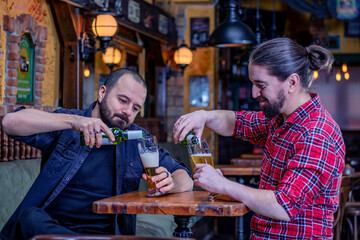 Two male friends drinking in the pub. Pouring beer from the bottle in tall glasses. Back to normal life.
