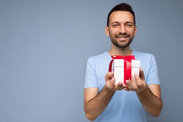 Photo shot of handsome positive smiling brunette unshaven young man isolated over blue background wall wearing blue t-shirt holding white gift box with red ribbon and looking at camera