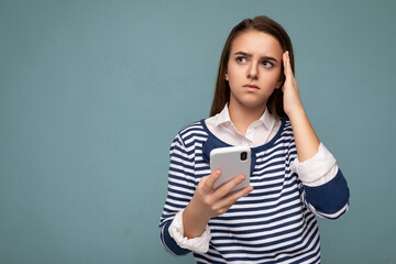 Naklejka premium Photo of thoughtful concentrated attractive young brunette girl wearing striped longsleev standing isolated over blue background surfing on the internet via phone looking up and thinking