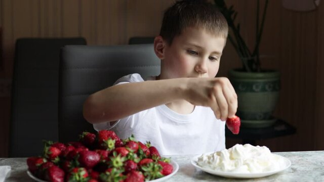 Boy eating strawberries with cream sitting near kitchen table at home. Schoolboy eating healthy organic food, fresh berries. Happy Childhood and Organic Products