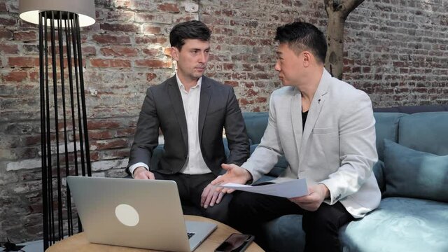 European Asian business partners discuss a commercial proposal in the meeting room shaking hands. Multinational male colleagues sitting on sofa holding contract documents negotiating business deal.