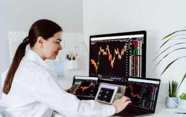 Young female cryptocurrency trader sitting at desk looking at multiple screens monitoring stock financial market charts. Business woman investor analyst analyze currency growth graph.
