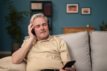 Elderly man sitting on the couch in the living room, listening to music from his phone on headphones in the evening. Tired boss relaxes on couch listening to songs from smartphone.