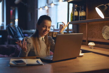 Young beautiful woman sits at a wooden table in a cafe indoors and works on a laptop. Conception of remote work and freelance work.