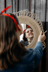 Obraz na płótnie Canvas The brunette artist is reflected in a round mirror, which she holds in her hands along with a brush. She looks at herself in reflection and smiles cheerfully on her cheeks with white traces of paint.