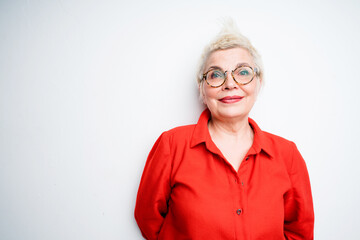 Beautiful cute elderly woman in glasses and in a red shirt smiling leaning against the wall