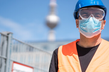 Occupational safety and protection against adverse conditions at work. Construction worker wearing hardhat, reflective vest and protective surgical mask. On background out of focused Berlin tv tower