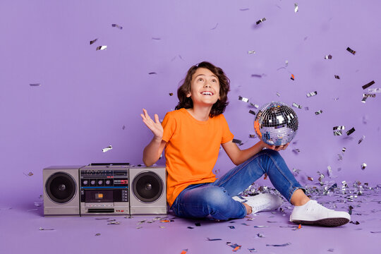 Full length body size photo boy sitting on boom box keeping disco ball with flying confetti isolated purple color background
