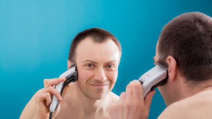 Mature man is cutting his hair by electric hair clipper in front of the mirror. - obrazy, fototapety, plakaty