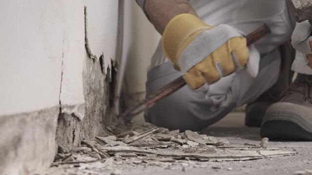 Construction worker hands with gloves working with hammer and chisel to remove old plaster from wall for house renovation, close up with rubble and tools