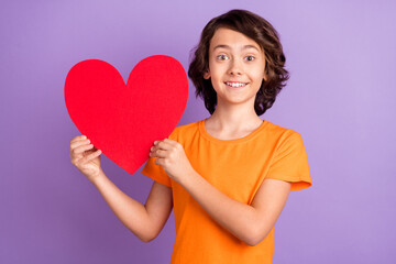 Fototapeta na wymiar Portrait of attractive cheerful boy holding in hands big paper heart isolated over violet purple color background