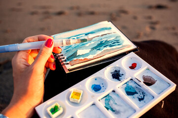 Plain air on a beach. Portable palette with watercolor, waterbrush and sketchbook on knees of a girl on a black sand seashore. Seaside leisures. Painting landscape outdoors for fun and health.