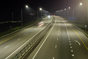  High-speed suburban highway in the light of streetlights. There are yellow and white markings on the asphalt. There are signs on the side of the road © VeNN