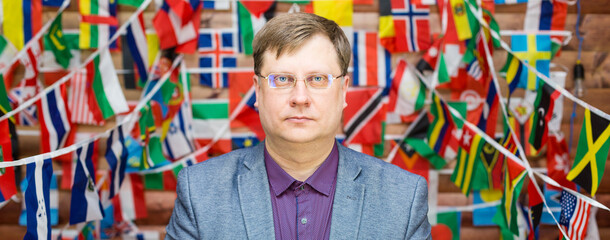 A strict man in glasses stands against the background of flags in the room.