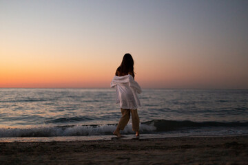 A slender girl in white clothes walks along the beach against the background of the sea. Dawn on the sea. A sportive girl at dawn runs along the embankment of the sea.