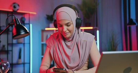 Pretty smiling happy relaxed 30-aged arabic lady in light-violet hijab in headphones listening nice music in beautifully decorated room with colourful lighting - Powered by Adobe