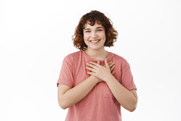 Thankful girl appreciate nice gesture, holding hands on heart, smiling grateful, express gratitude, being heartfelt and touched, standing in t-shirt over white background