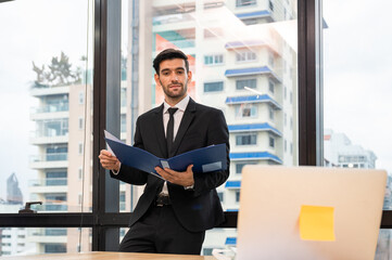Fototapeta premium Caucasian executive businessman standing holding document folder looking through laptop in the office