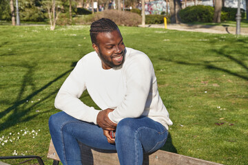 Obraz premium Happy African American man sitting in a park wearing jeans, white sweater. Lifestyle concept