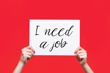 White sheet poster with the slogan I need a job in female hands isolated on a color red background. Board, blank, template, mockup, layout. Dismissal of workers. Job Search. Unemployed