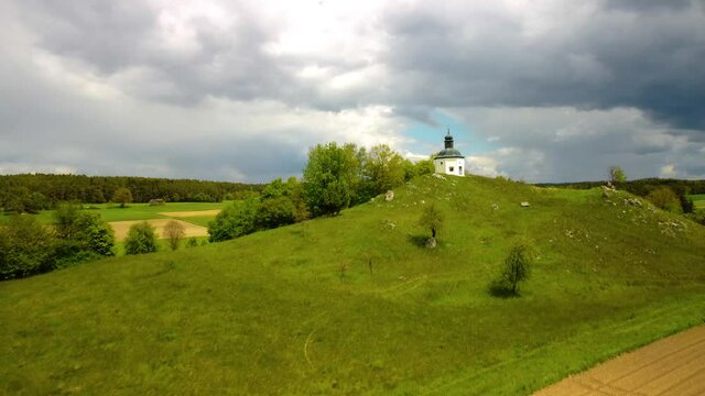 Drone flying over open fields and small chapel on a rocky hill with perfect view over typical Bavarian rural landscape on sunny day