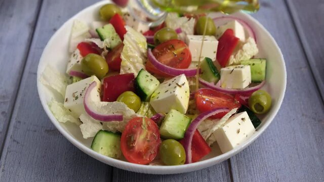 Pouring Olive oil on Greek salad. Traditional Greek dish. Healthy vegetarian food. Fresh vegetables and feta cheese in a white plate. Close-up.