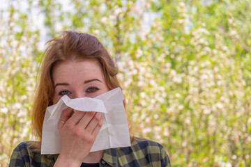 Season allergy to flowering plants pollen. Young woman with paper handkerchief in hand covering her nose in garden. Teen girl sneezing against blossoming trees. Seasonal allergy prevention - obrazy, fototapety, plakaty