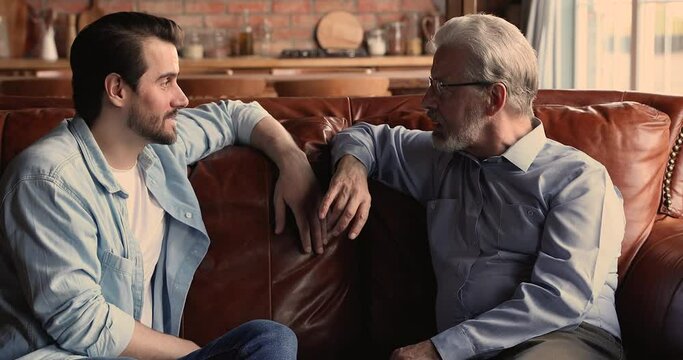 Older man his grown up millennial son sit on sofa and talking, share news enjoy moments together having pleasant conversation and arm friendly harmonic relations between two diverse generation concept
